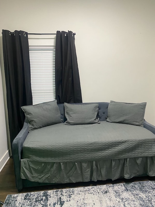 A bed with a grey quilt and pillows, a window with blinds, and a dark curtain.