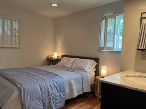 Ein Schlafzimmer mit Bett, Nachttischlampen, einem Nachttisch, einem Fenster mit Jalousien und einem Badezimmer mit Waschbecken.