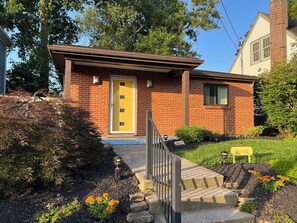 A brick house with a yellow door, a front porch, and a small garden with flowers and shrubs. - Hyde Park "Rail Trail" Entire House (Cincinnati)