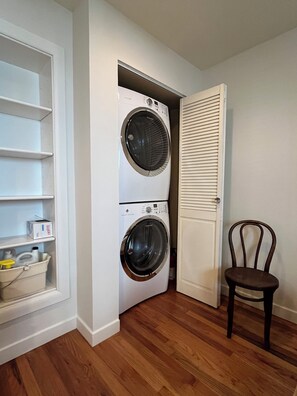 A laundry room with two stacked front-loading washing machines, a chair, and a shelf with cleaning supplies. - Hyde Park "Rail Trail" Entire House (Cincinnati)