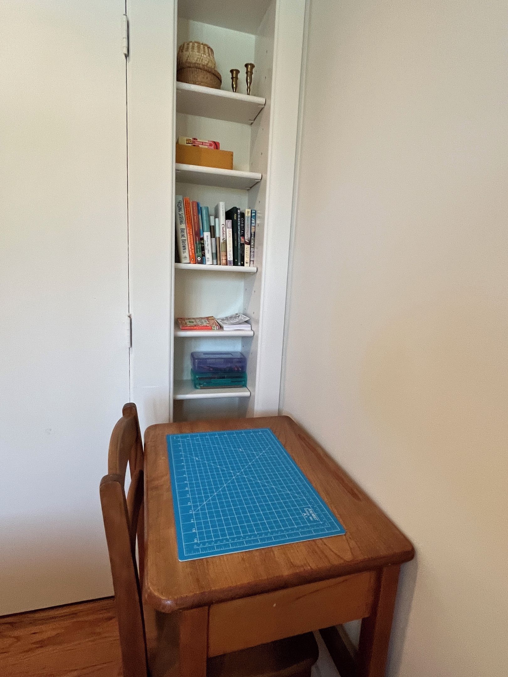 Una silla de madera con un tapete de corte azul sobre una mesa, un estante blanco con libros y recipientes, y una puerta blanca.
