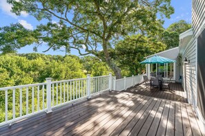 Eine Holzterrasse mit weißer Geländer, ein überdachter Sitzbereich inklusive Tisch und Stühlen sowie Blick auf ein bewaldetes Gebiet.