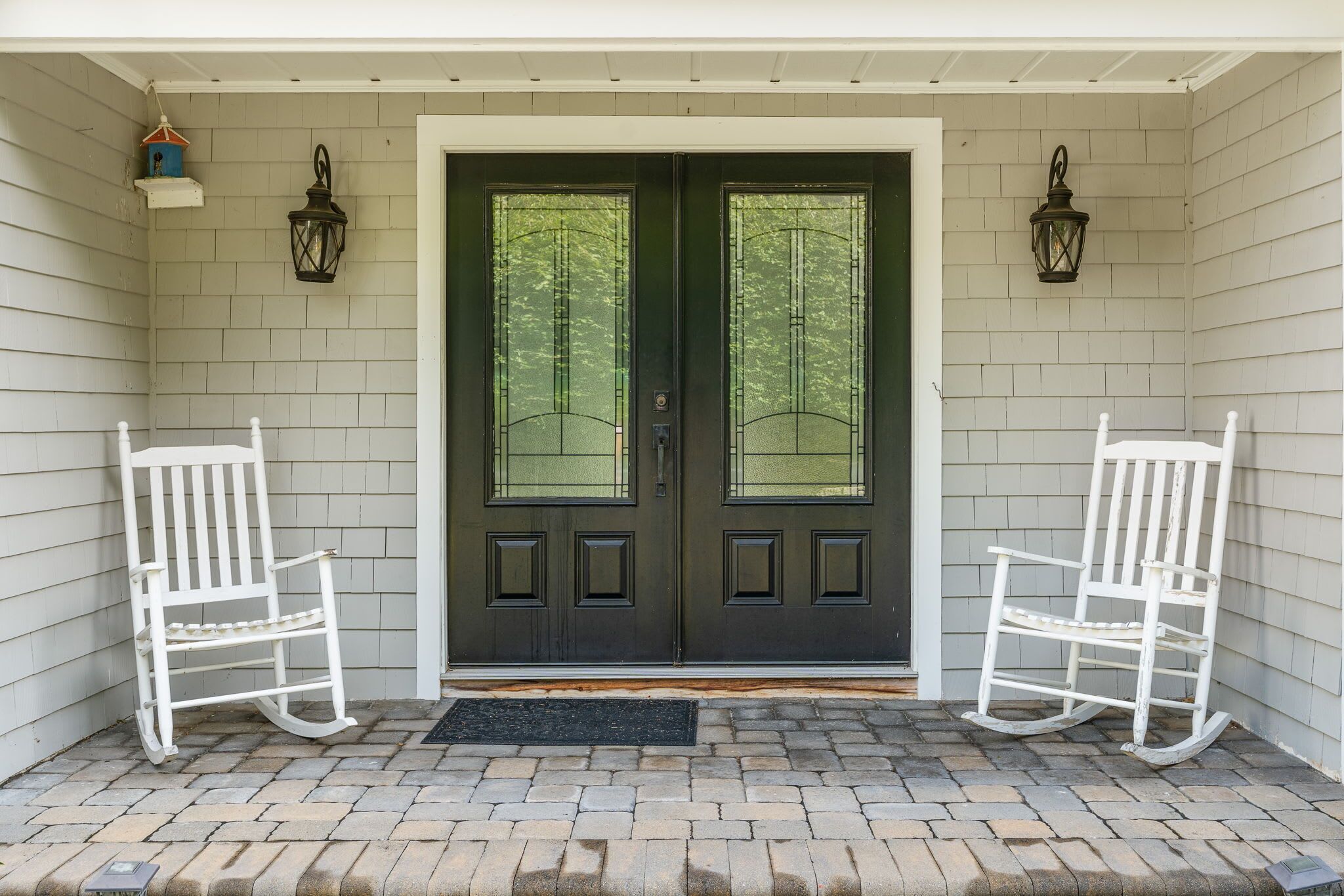 A front door with two white rocking chairs on either side, a black door with a window, and a brick-paved entrance.