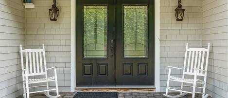 A front door with two white rocking chairs on either side, a black door with a window, and a brick-paved entrance.