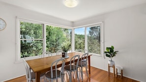 A dining area with a wooden table and chairs, large windows with a view of greenery, and a clock on the wall. - Behind the Dunes - Point Lonsdale (Point Lonsdale)