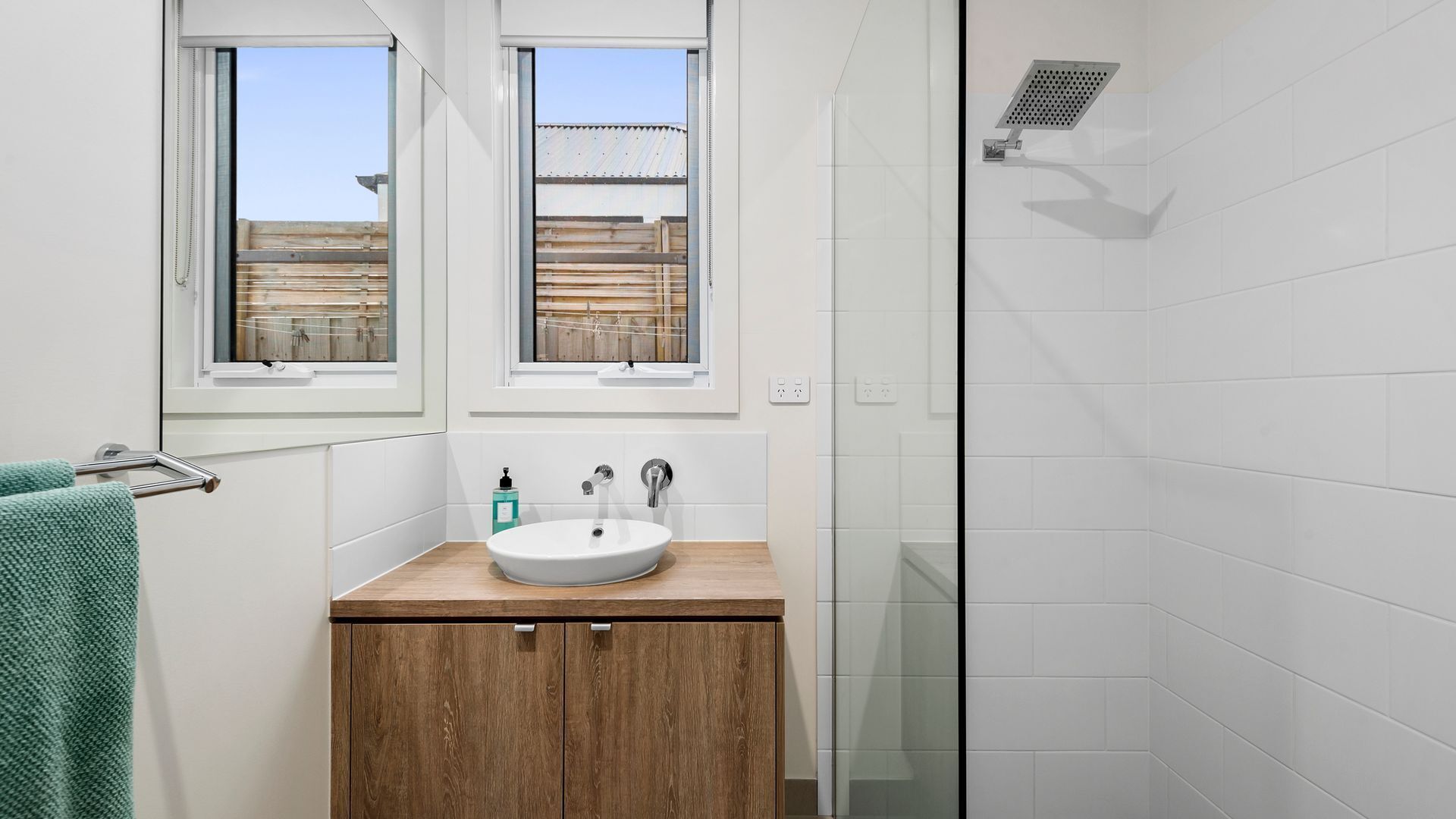 A bathroom with a wooden vanity, a sink, a glass shower enclosure, and a view of a building outside through a window.