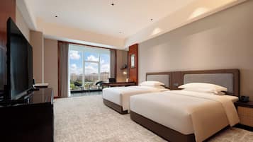 A hotel room with two beds, a desk, a TV, and a large window with a city view.