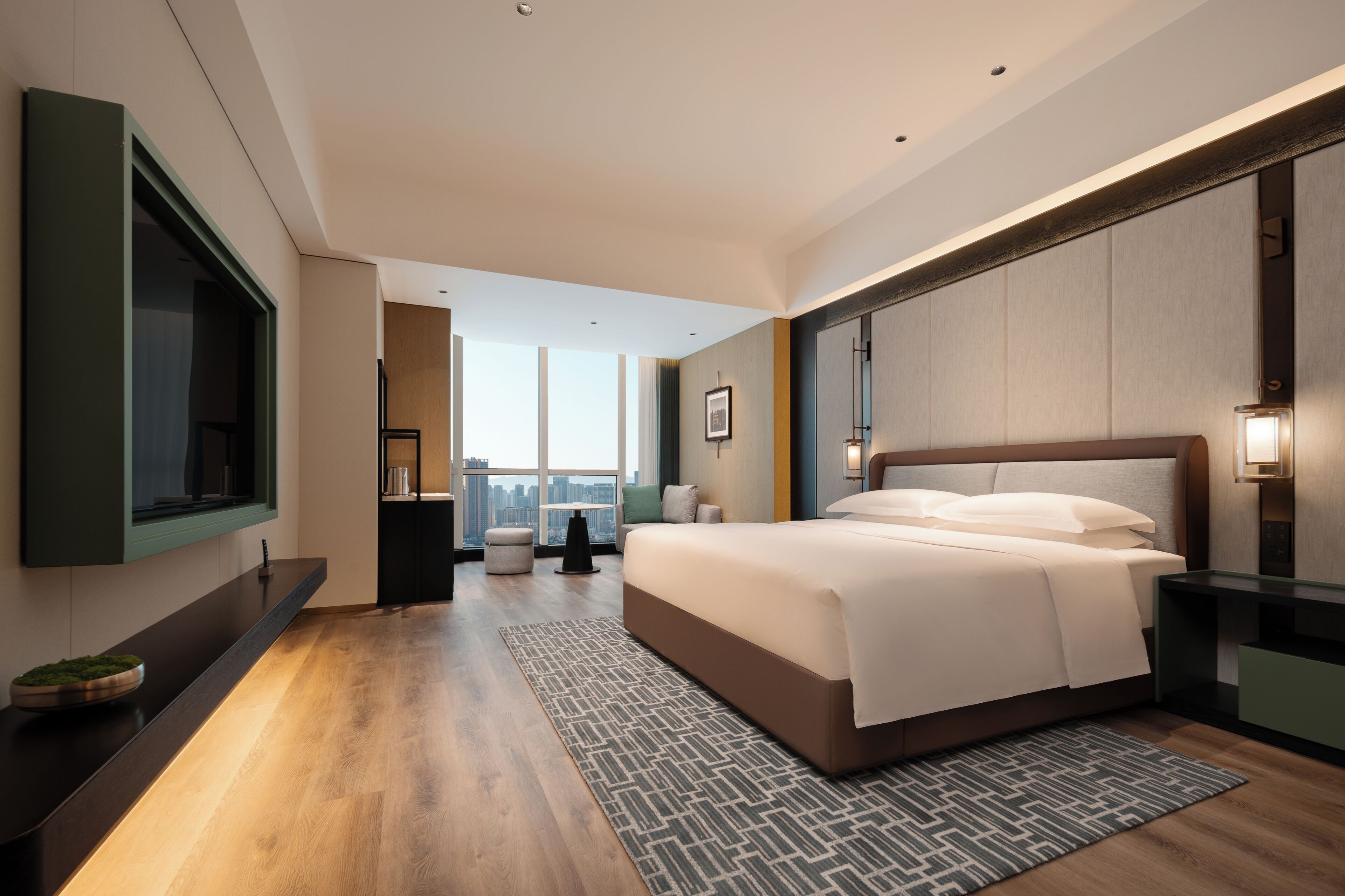 A modern hotel room with a large bed, a television, a city view, and a small seating area.