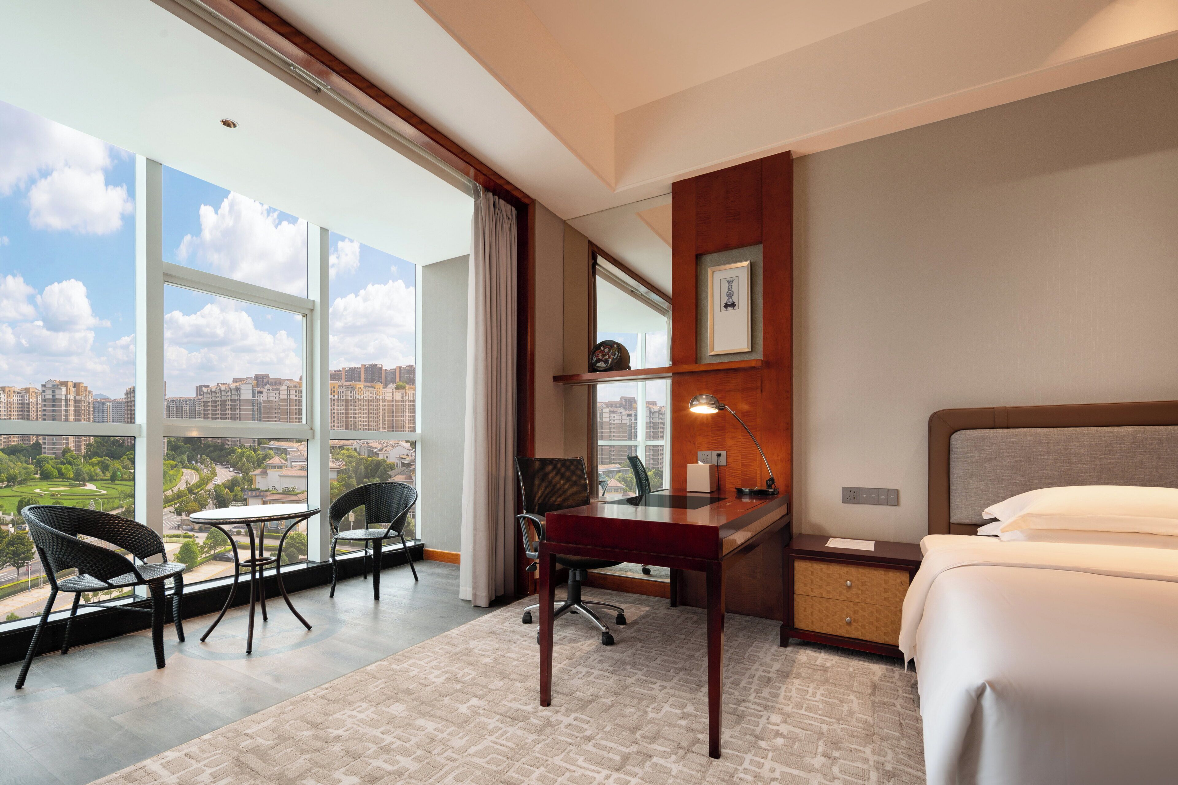 A hotel room with a large window, a desk, a chair, a bed, and a nightstand.