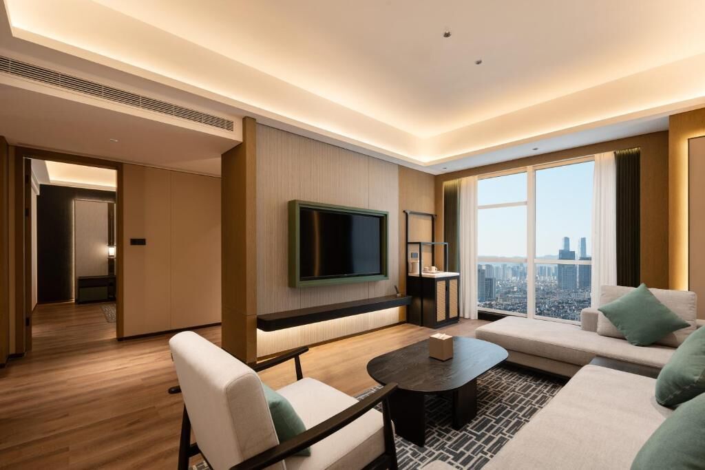 Habitación de hotel moderna con un ventanal grande, televisor de pantalla plana, sofá, mesa de centro y vista panorámica de la ciudad.