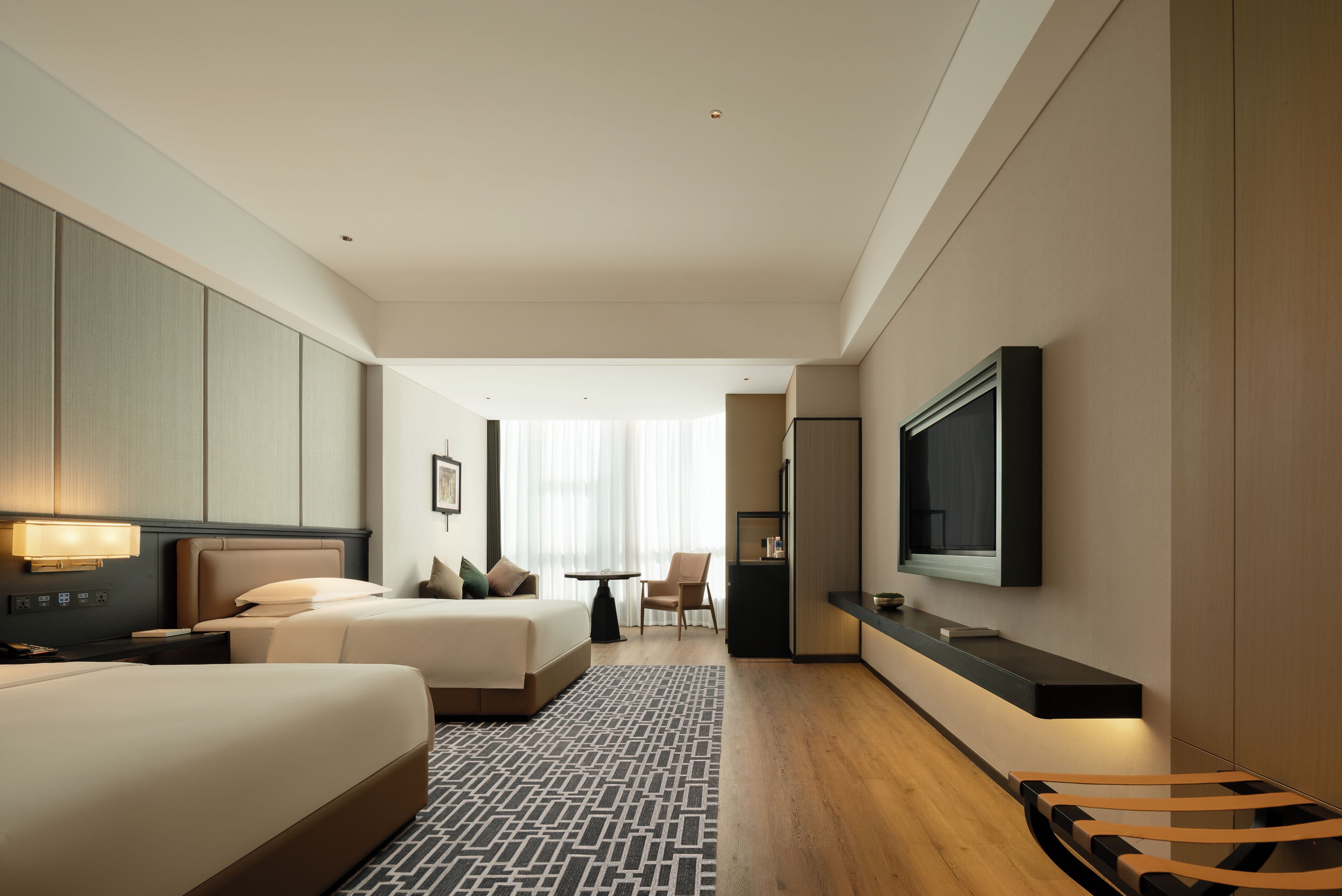 A hotel room with a bed, bedside table, lamp, TV, and a small seating area with a chair and table.