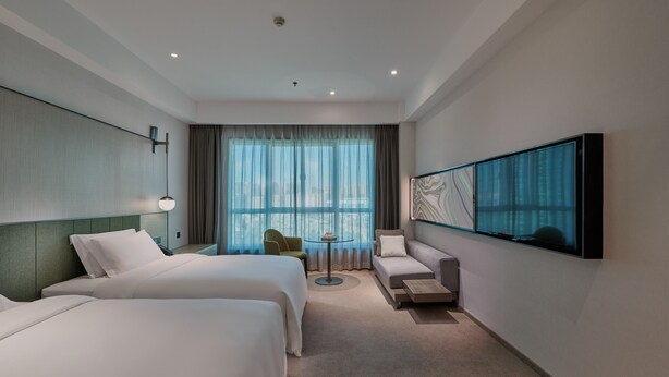 Habitación de hotel con dos camas, un sofá, un televisor y vista a la ciudad a través de amplias ventanas.
