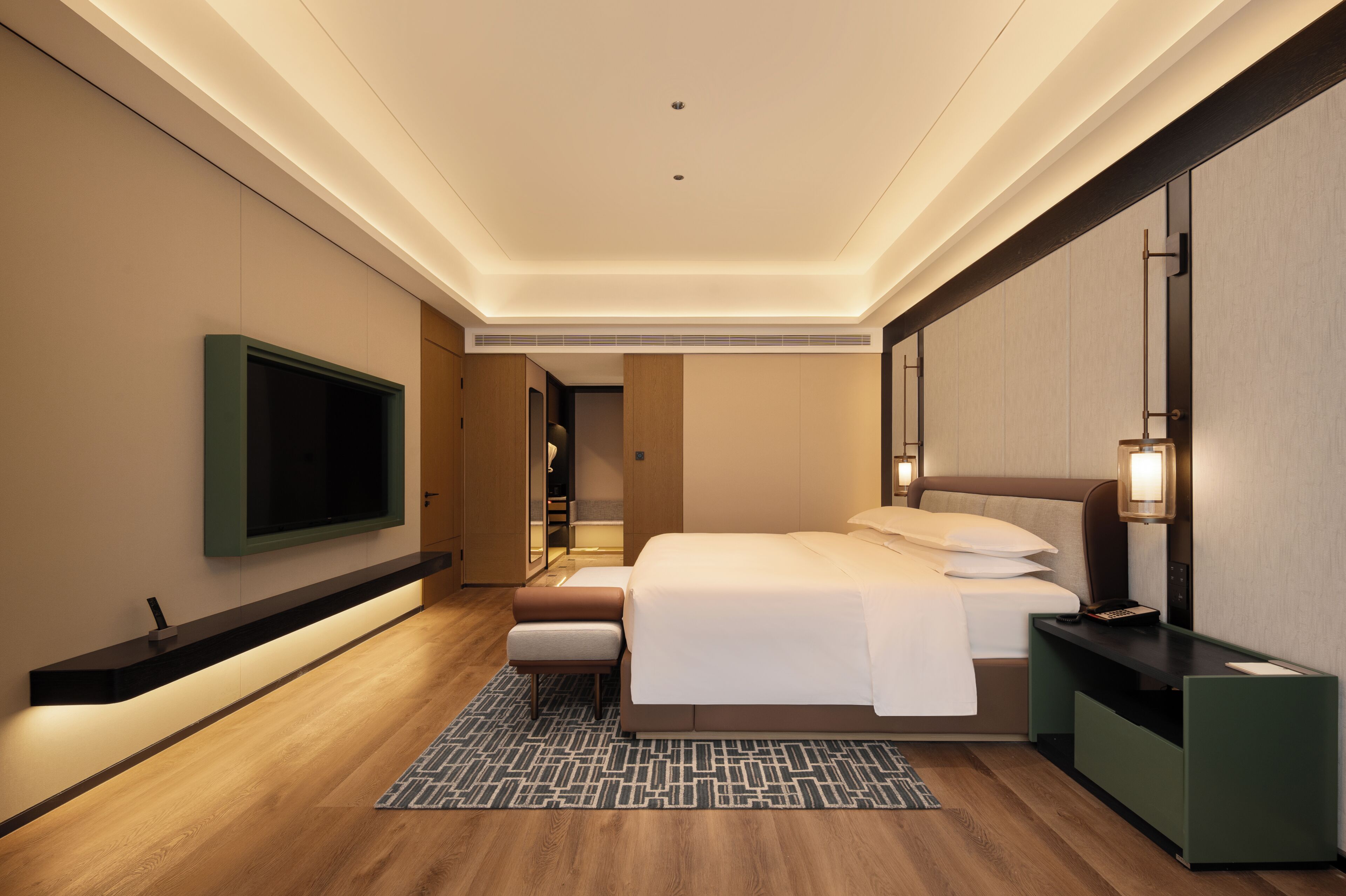 A modern hotel room with a large bed, a flat-screen TV mounted on the wall, a bedside table with a lamp, and a built-in wardrobe.