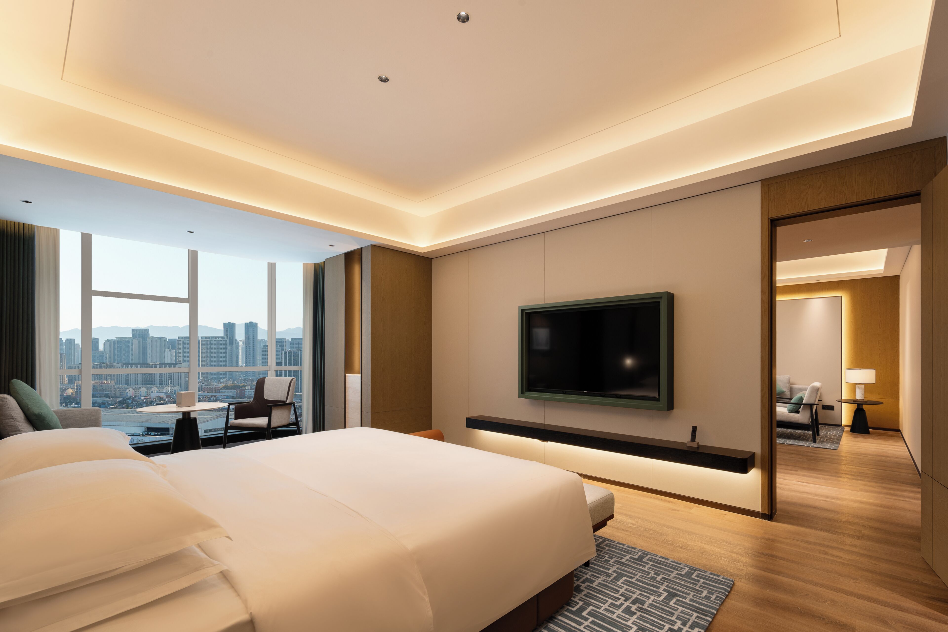 A modern hotel room with a large bed, a flat-screen TV mounted on the wall, and a view of the cityscape through large windows.