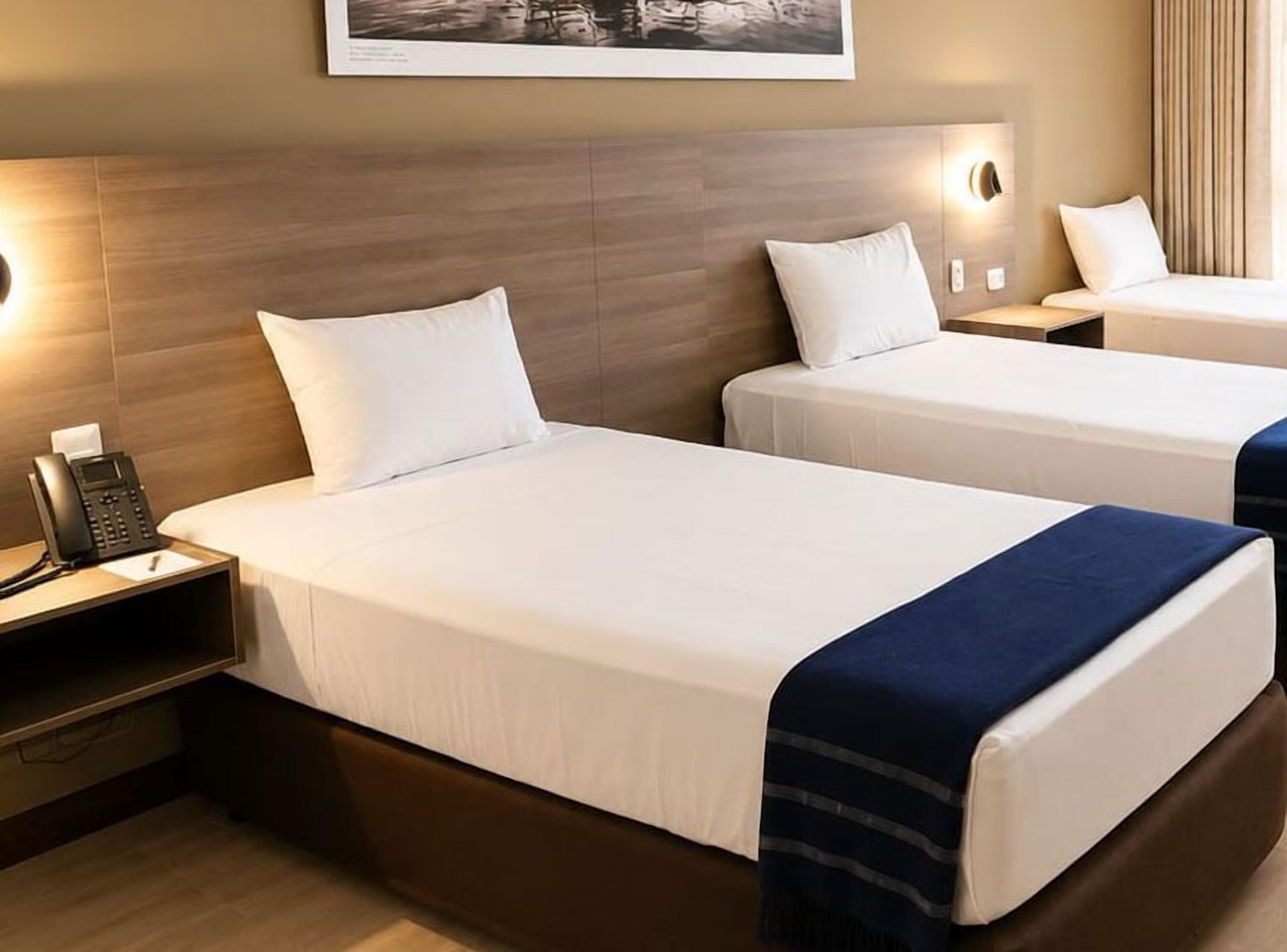 A hotel room with two beds, a bedside phone, and a wall-mounted light fixture.