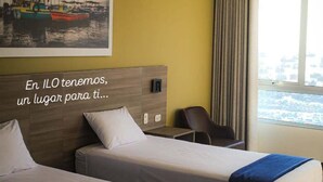 A hotel room with two beds, a headboard with a message, a painting of boats, a window with curtains, and a view of the city. - Hotel Costa del Sur (Ilo)