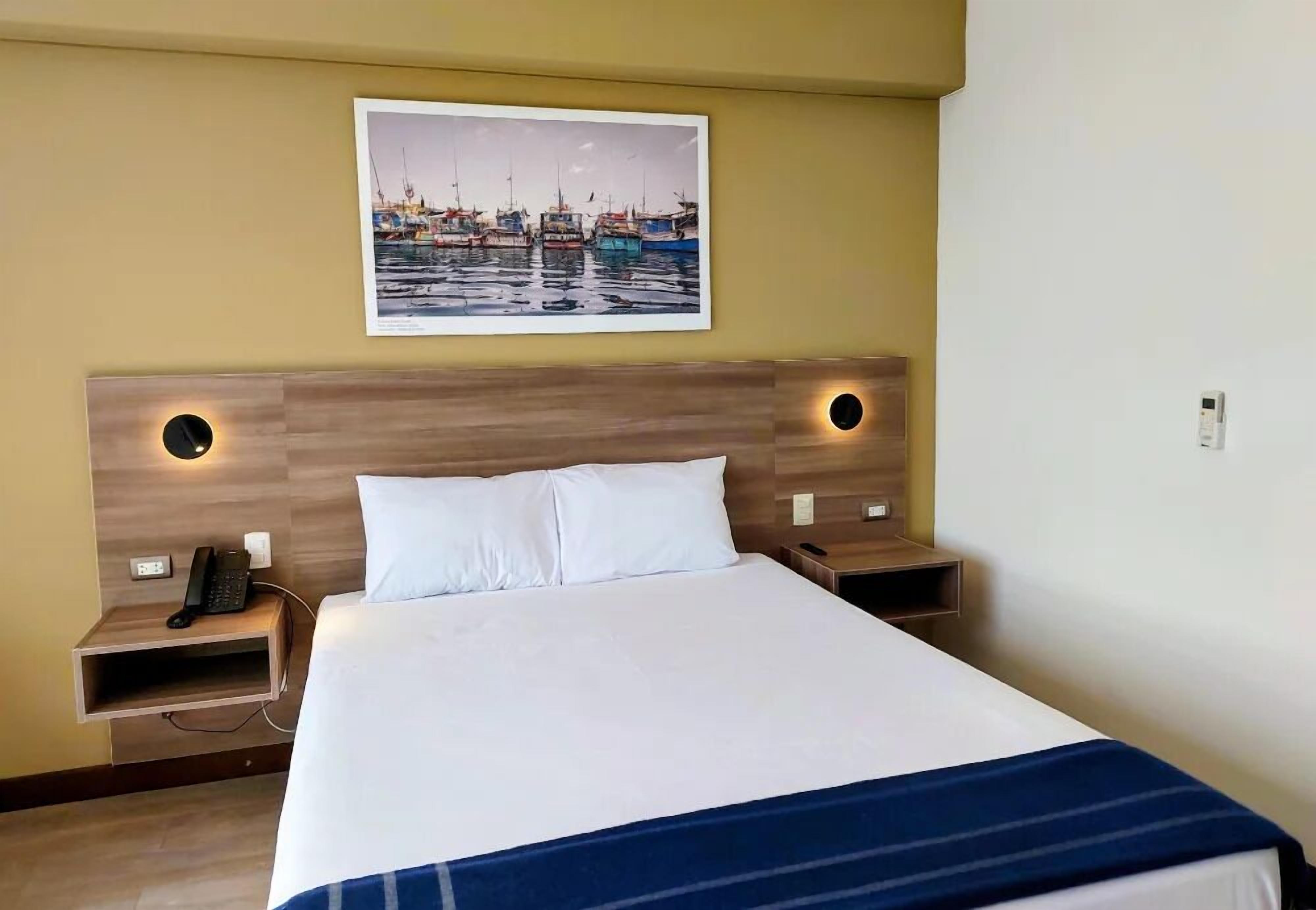 A hotel room with a large bed, two bedside tables, a wall-mounted telephone, and a framed picture of a harbor.