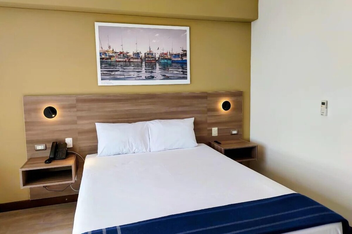 A hotel room with a large bed, two bedside tables, a wall-mounted telephone, and a framed picture of a harbor.