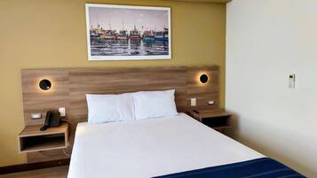 A hotel room with a large bed, two bedside tables, a wall-mounted telephone, and a framed picture of a harbor.