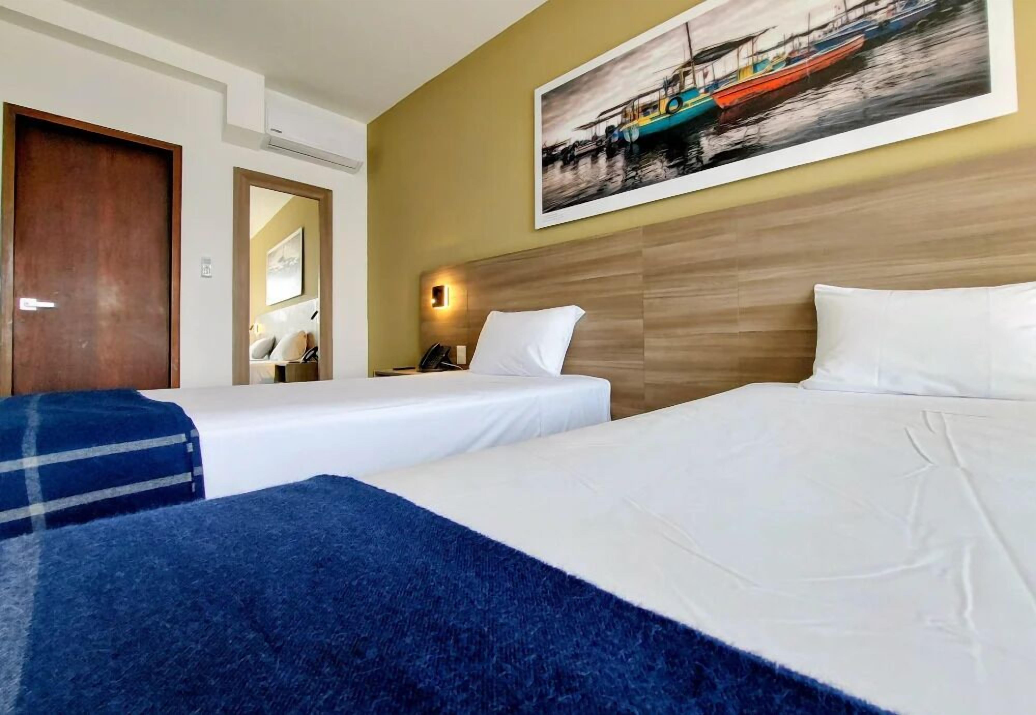 Une chambre d’hôtel avec deux lits, une tête de lit en bois, un tableau représentant des bateaux et un miroir.