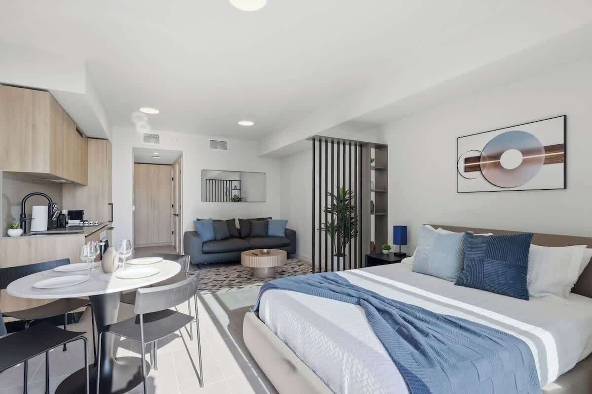 A modern hotel room with a bed, a dining table, and a kitchen area.