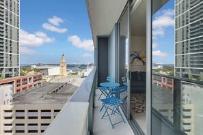 Un balcon avec une table et des chaises, d’où l’on peut admirer un paysage urbain avec des immeubles et une tour située au loin.