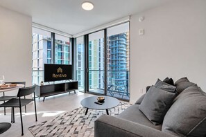 Ein modernes Wohnzimmer mit einem Flachbildfernseher, einer Couch, einem Esstisch und Blick auf hohe Gebäude durch große Fenster.