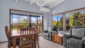 A room with a dining table, chairs, a sofa, and a small bedside table. There are large windows providing a view of trees and a clear sky. - Lamont House Bermagui - Lamont Street 104 (Bermagui)