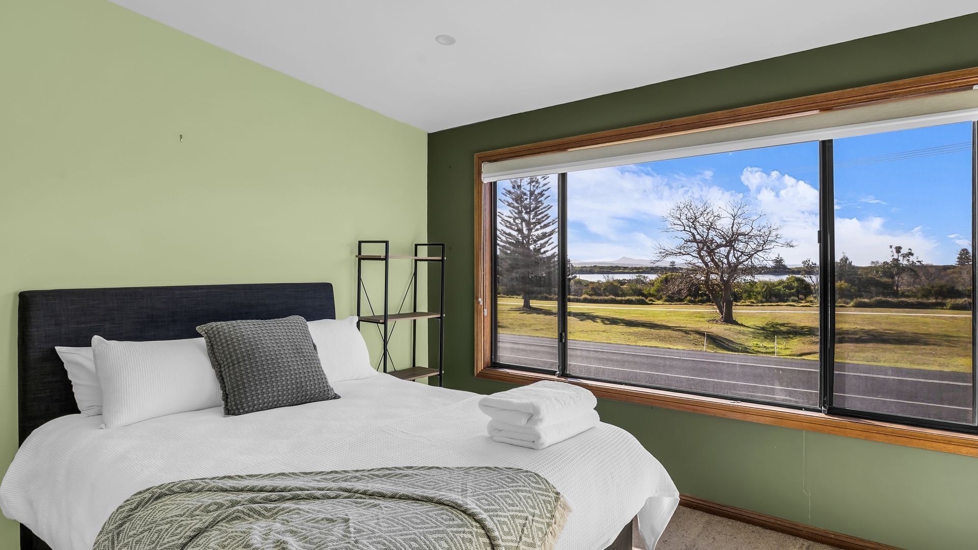 A bedroom with a large window, a bed with white bedding, and a view of a road and trees.