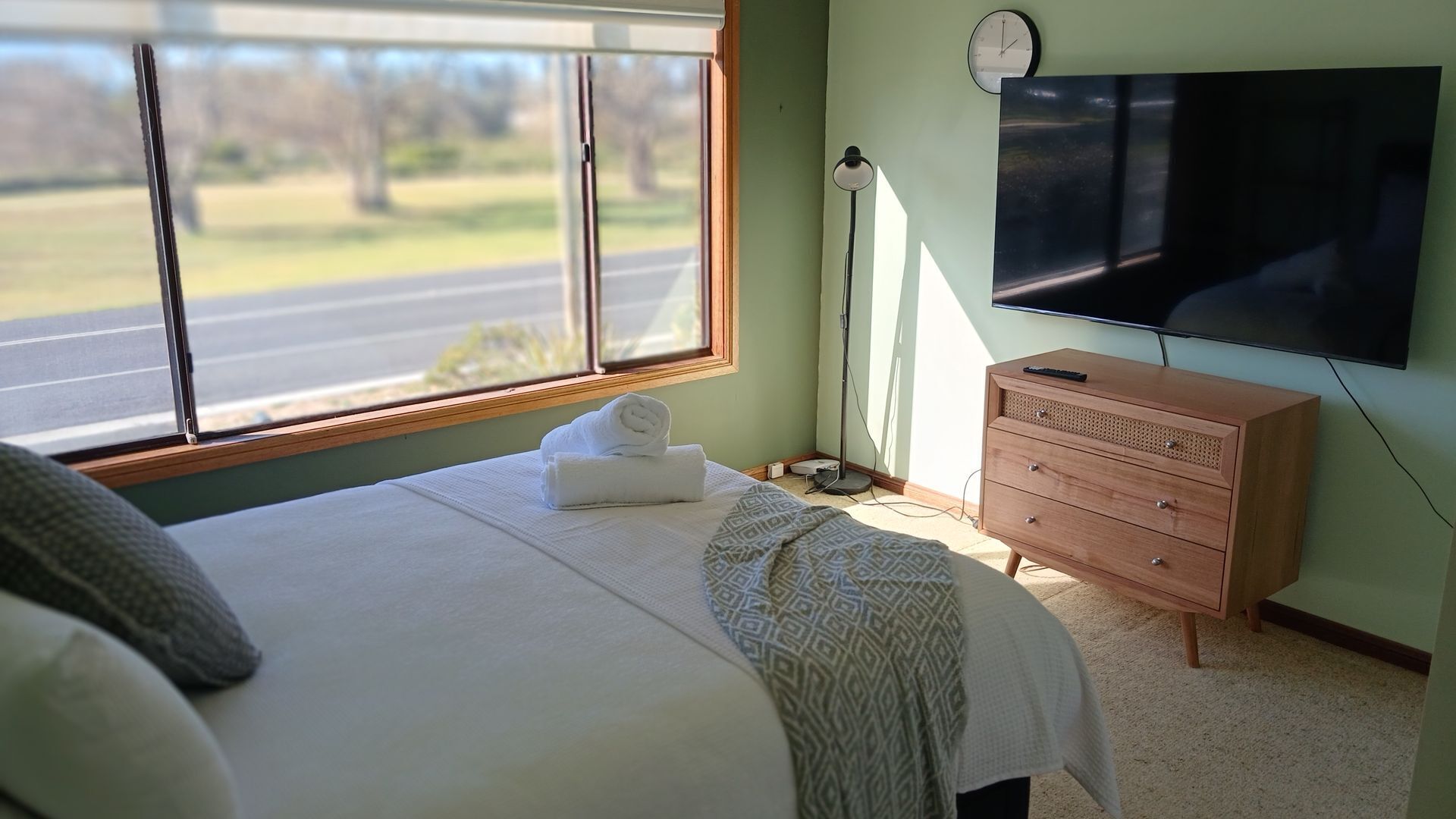 Ein Schlafzimmer mit Bett, Kommode, Fernseher und Fenster mit Blick auf eine Straße und Bäume.