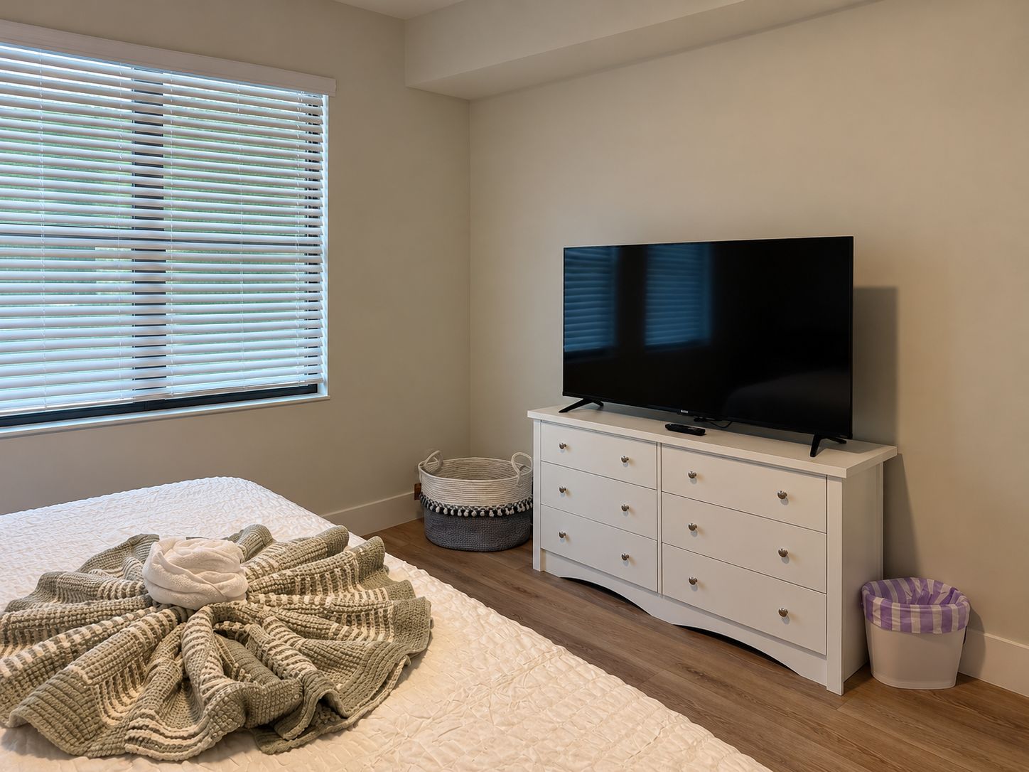 Una camera da letto con un letto, una televisione su un comò, una finestra con persiane e un cestino.