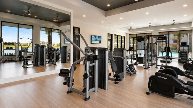 Een sportschool met diverse fitnessapparaten, grote ramen en een plafondventilator.