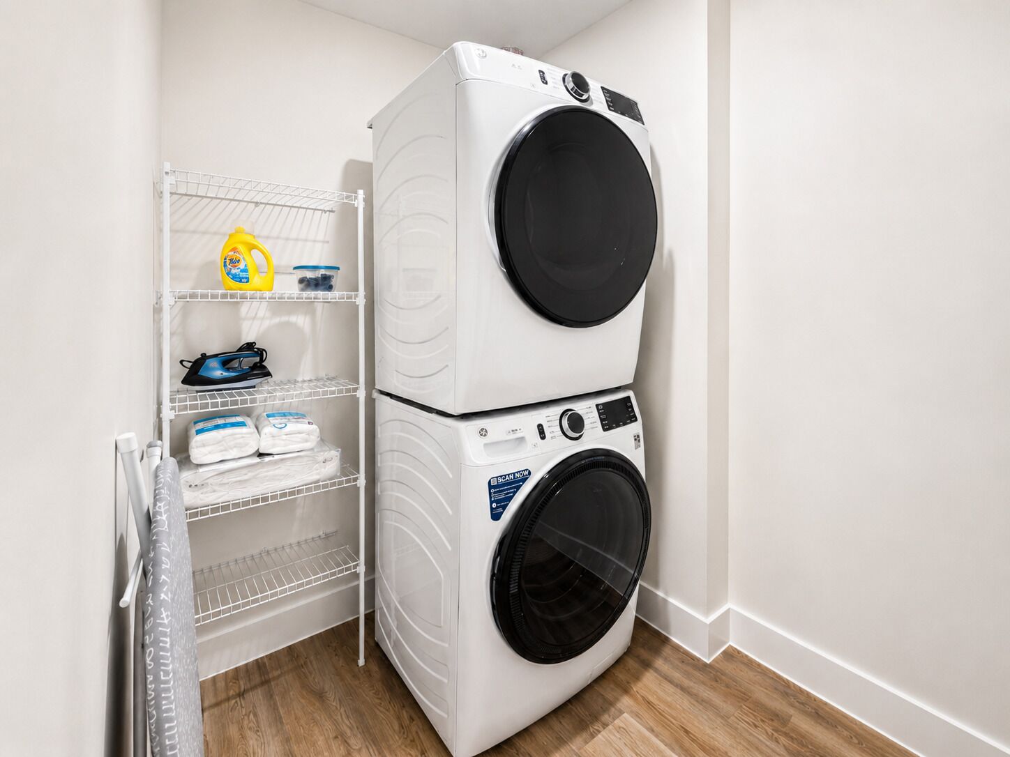 Un cuarto de lavandería con dos lavadoras de carga frontal apiladas y un estante metálico con productos de limpieza.