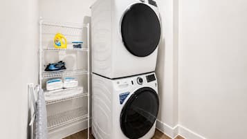 Un cuarto de lavandería con dos lavadoras de carga frontal apiladas y un estante metálico con productos de limpieza.