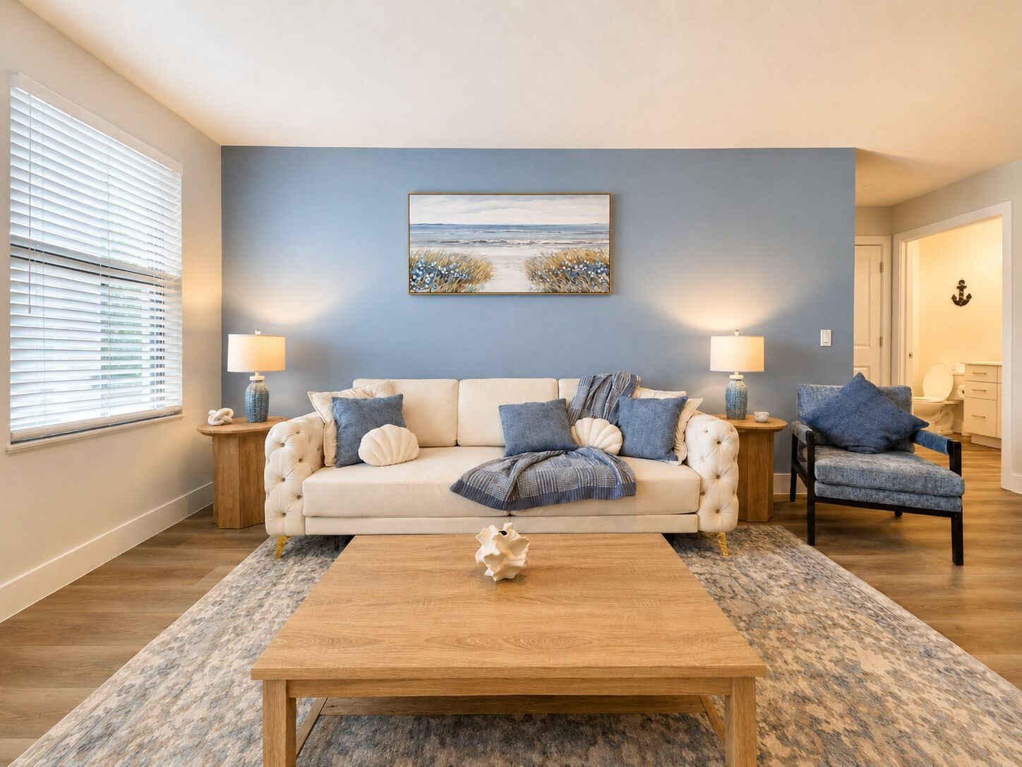 Un salon avec un canapé beige, deux tables d’appoint avec des lampes, une table basse et un tableau représentant une scène de plage accroché au mur.