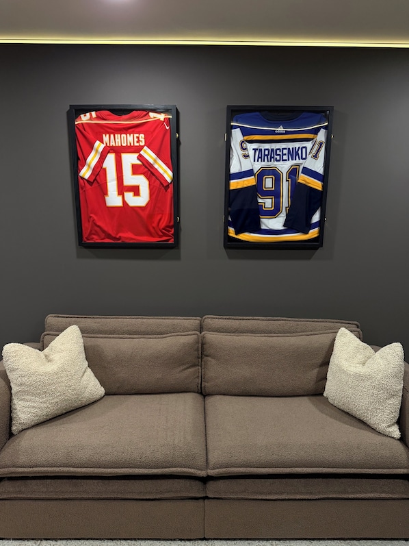 Two framed sports jerseys on a wall above a brown sofa with two pillows.