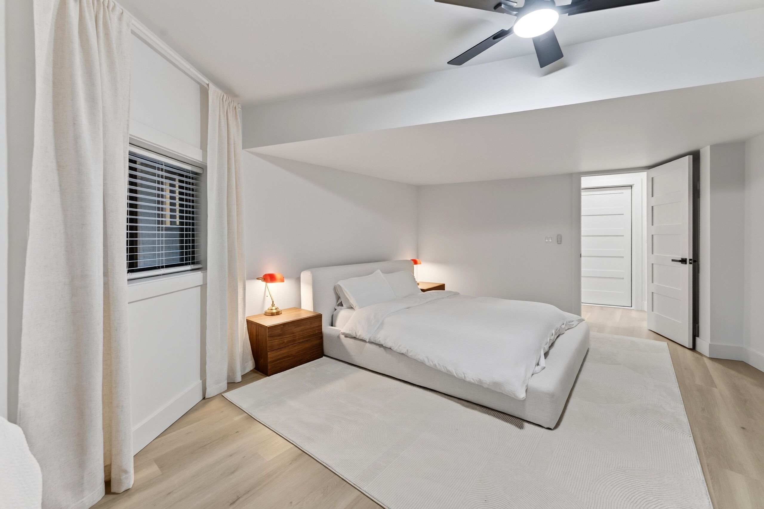 Un dormitorio con cama, mesitas de noche, ventilador de techo, ventana con persianas y puerta.