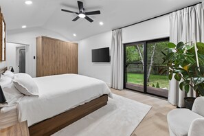 Ein modernes Schlafzimmer mit einem großen Bett, einem Deckenventilator, einem Fernseher und Blick auf einen Garten durch eine Schiebetür.