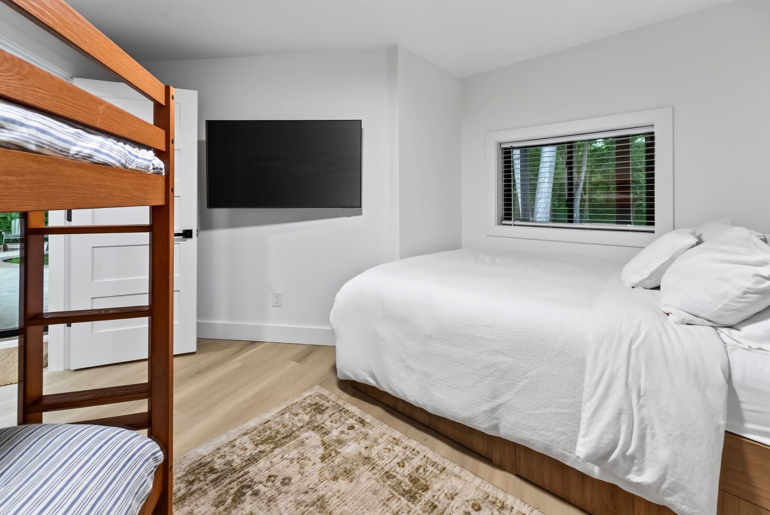 A bedroom with a bunk bed, a flat-screen TV mounted on the wall, a window with blinds, and a bed with white bedding.