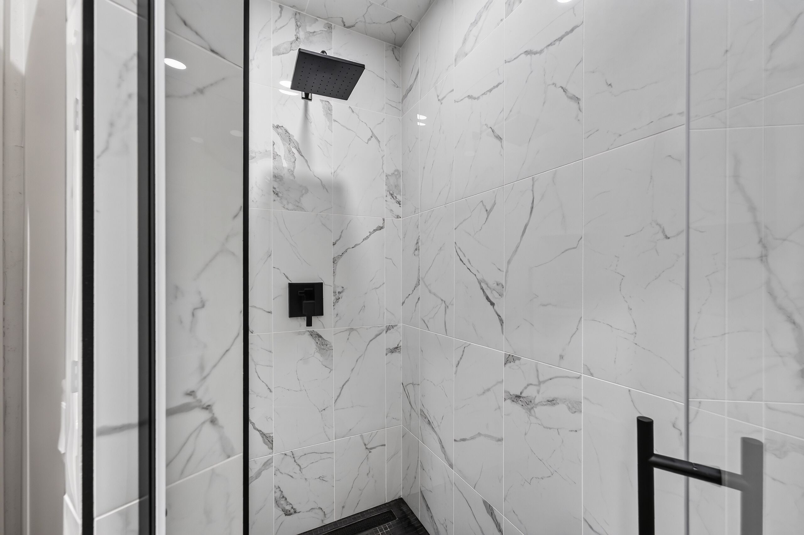 A modern bathroom with white marble tiles, a glass shower enclosure, and a black towel rack.