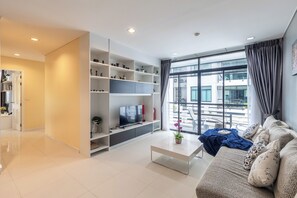 A modern living room with a flat-screen TV, a sofa, and a coffee table. There is a large window with curtains, and a bookshelf filled with decorative items. - The Regent Kamala Condominium by PSR (Tambon Kamala)