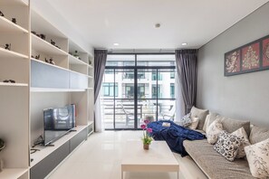 Una sala moderna con un sofá, una mesa de centro y un televisor sobre un mueble bajo. Hay estantes con objetos decorativos y un ventanal con cortinas.
