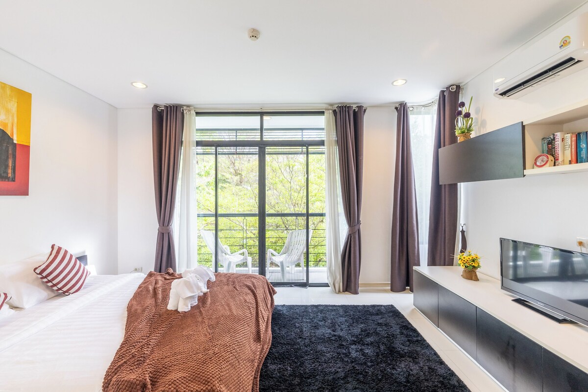 A modern hotel room with a bed, a TV, a large window with curtains, and a view of greenery outside.