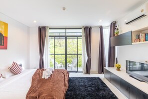 A modern hotel room with a bed, a TV, a large window with curtains, and a view of greenery outside. - The Regent Kamala Condominium by PSR (Tambon Kamala)