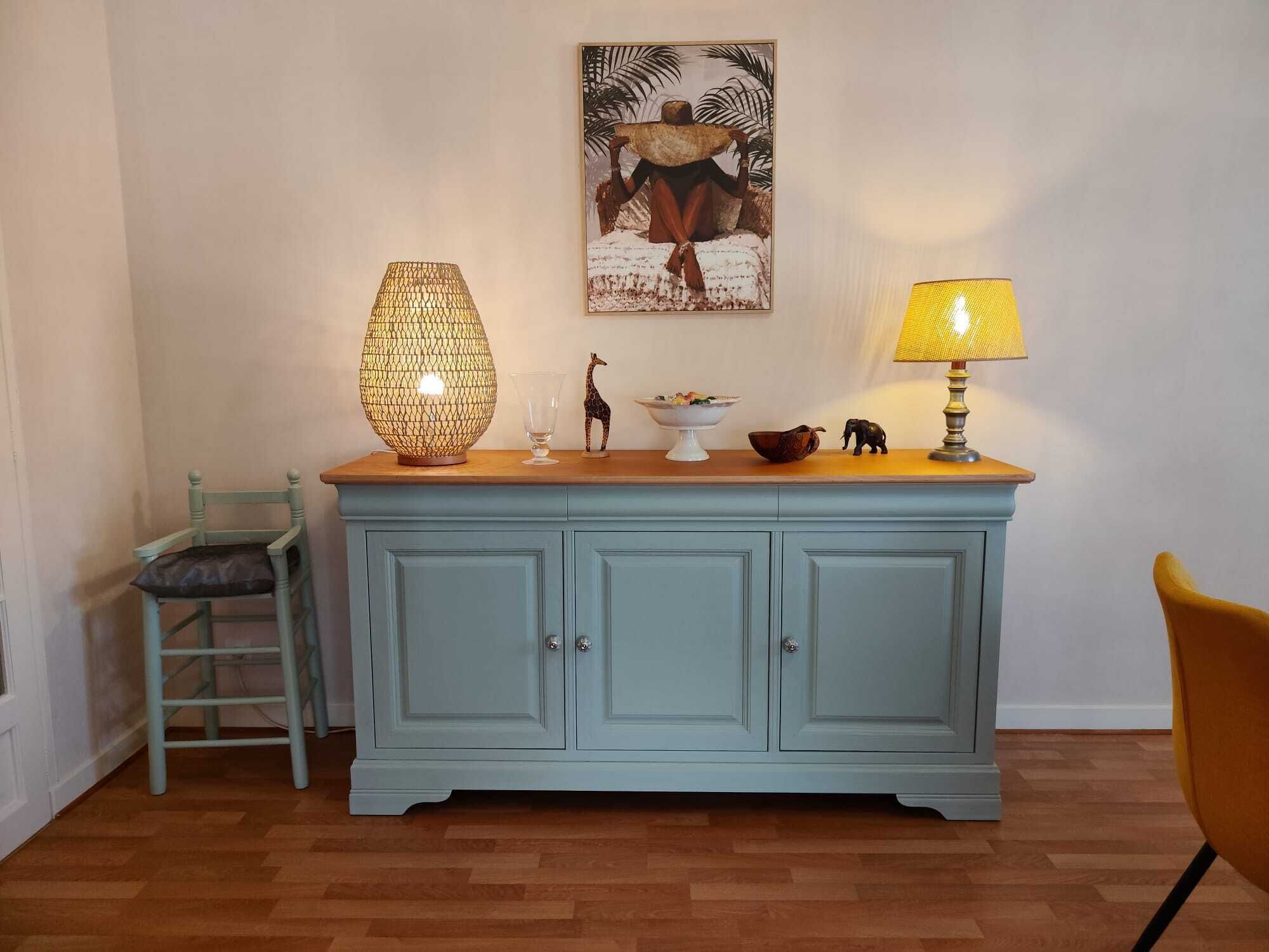 A light blue wooden cabinet with a glass top, a wicker lamp, a small giraffe figurine, a bowl of fruit, and a lamp with a yellow shade.