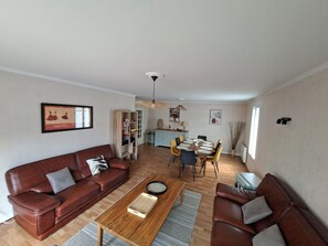 Een woonkamer met een bruine leren bank, een houten salontafel, en een eethoek met een ronde tafel en stoelen.
