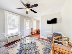 A living room with a ceiling fan, a flat-screen TV mounted on the wall, a glass coffee table, and a patterned rug.