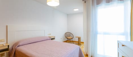 A bedroom with a bed, bedside tables, a chair, a small table, a lamp, and a window with curtains.