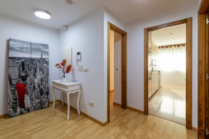 A modern interior with a wooden floor, a white console table with a vase of red flowers, a cityscape painting, and a view into a bright kitchen area. - Volvoreta Valdoviño Apartment (Valdoviño)