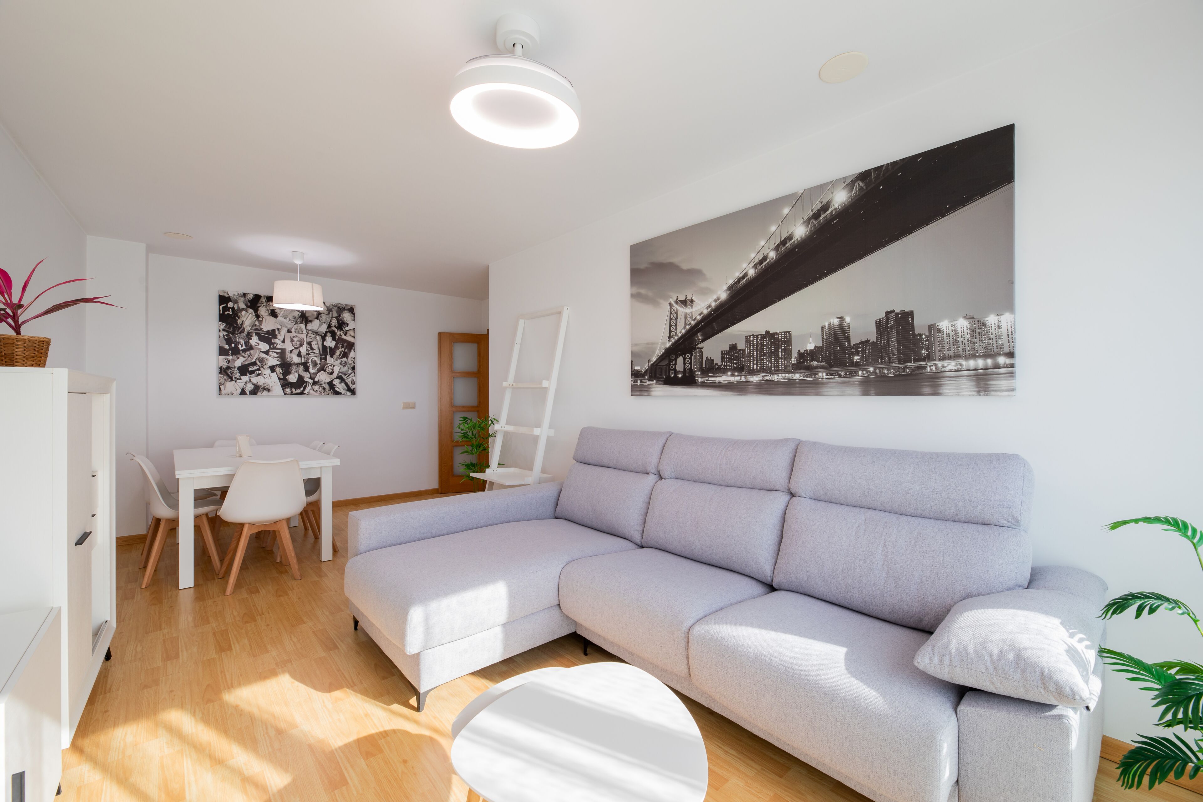 Ein modernes Wohnzimmer mit einem grauen Ecksofa, einem Esstisch mit Stühlen, einem weißen Couchtisch und einem Stadtlandschaftsfoto an der Wand.